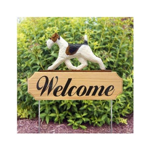 May include: A wooden sign with the word "Welcome" in elegant script. A carved dog figurine in white, brown, and black is mounted above the sign. The sign is on metal stakes, set against a backdrop of green foliage.