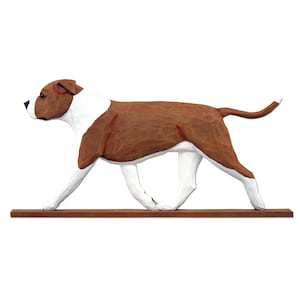 May include: A wooden sculpture of a dog in a walking pose. The dog has a brown body with white markings on its chest, legs, and head. It is mounted on a brown wooden base. The dog's tail is slightly curved.