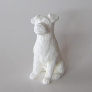 May include: A white, seated dog figurine with a textured surface. The dog has upright ears and a detailed snout, with its front paws resting on the ground. The sculpture is a solid, off-white color.