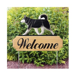 May include: A wooden welcome sign featuring a black and white dog silhouette. The sign has the word "Welcome" in black script. The sign is mounted on metal stakes and is set against a backdrop of green foliage.