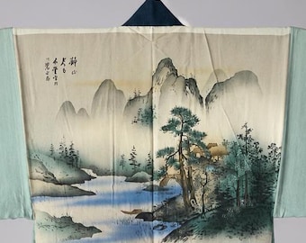 May include: A kimono featuring a landscape painting on the back panel, depicting mountains, trees, and a river in shades of green, blue, and cream. The garment has wide sleeves with dark blue cuffs and a solid dark blue lower section with text.