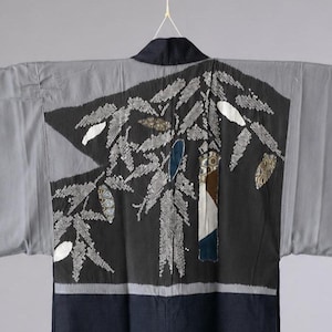 Puede incluir: Un kimono gris y azul marino oscuro con mangas anchas y cuello oscuro. El panel trasero muestra un gran elemento gráfico central negro decorado con patrones de hojas blancas, intercalado con pequeños parches de tela texturizada.