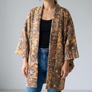 Op de afbeelding: Beige kimono-jas met een bloemenpatroon in rood, blauw en groen. De jas heeft wijde mouwen en een losse pasvorm. De persoon draagt een zwarte top en een blauwe jeans. De jas is een uniek kledingstuk.