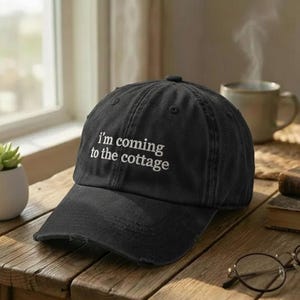 May include: Black baseball cap with the embroidered text "I'm coming to the cottage." The cap has a distressed look and a curved brim. The hat is sitting on a wooden surface.