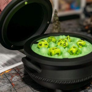 May include: A black cauldron-shaped dice box with a hinged lid, filled with green foam and yellow dice. The dice are numbered with white markings. The cauldron has three small feet and two side handles.