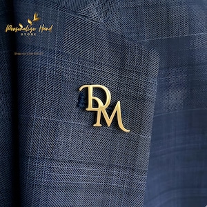 Könnte beinhalten: Ein goldfarbener Ansteckpin mit den Initialen "DM" ist an einem marineblauen karierten Anzugsakko befestigt. Der Pin, in einer Schreibschrift, ist ein stilvolles Accessoire. Das Sakko hat ein dezentes Karomuster.