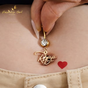 May include: A gold-colored belly button ring with a clear gemstone and the name "Reece" in a cursive font. A small red heart is next to the belly button. The image is of a person's torso.