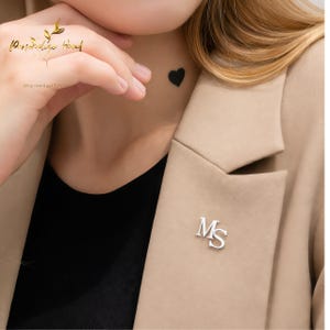 Puede incluir: Un broche plateado con las letras "MS" en una blazer color beige. La imagen también muestra una camiseta negra, un tatuaje de corazón negro y el texto "Personalize Hand".