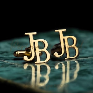 Op de afbeelding: Goudkleurige manchetknopen met de initialen "JB" in een vetgedrukte lettertype. De manchetknopen worden gepresenteerd op een reflecterend, blauwgroen oppervlak met een donkere achtergrond. De afbeelding toont de gepolijste afwerking en de weerspiegeling van de initialen.