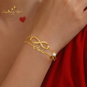 May include: A gold-coloured bracelet with an infinity symbol set with small, clear crystals. The bracelet includes a chain with the name "Angela" in cursive script and a heart-shaped charm. The image also shows the text "Personalize Hand Store".