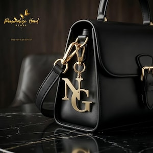 Puede incluir: Bolso de mano de cuero negro con herrajes dorados y un charm decorativo con las letras "NG". El bolso tiene un asa superior y una correa para el hombro. El texto "Personalize Hand Store" y "Shop now & get 50% Off" son visibles.