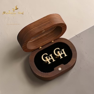 Op de afbeelding: Een open, ovaalvormige houten sieradendoos met een zwart fluwelen interieur. Binnenin zijn twee goudkleurige oorbellen zichtbaar, elk met de letters "GH". De doos staat tegen een neutrale achtergrond. De tekst "Personalized Hand" is zichtbaar.