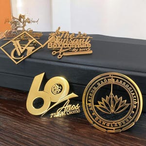 May include: Gold-colored metal pins and badges. Designs include a stylized "V" within a diamond, text "The Sunset Box/cottire", the number "60" with text "Aros", and a circular badge with text "Amateur Radio Association Bangladesh".