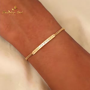 Puede incluir: Una pulsera dorada con una barra rectangular grabada con coordenadas: 36°53'47.4"N 76°10'42.9"W. La pulsera tiene un diseño de cadena fina. La imagen es un primer plano de la pulsera en una muñeca.