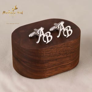 May include: A pair of silver cufflinks, each displaying the intertwined letters "AB", rests on a dark wooden box. The cufflinks are highly polished, reflecting light. The box has a smooth, oval shape. The image also contains the text "Personalized Hand".
