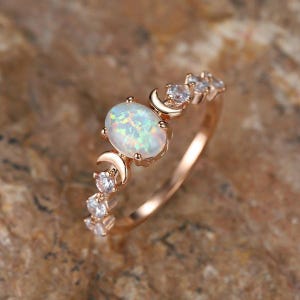 Puede incluir: Un anillo de oro rosa con una piedra de ópalo ovalada e iridiscente en el centro, flanqueada por dos motivos de media luna. La banda de metal está decorada con una serie de piedras transparentes de corte redondo a lo largo del aro.