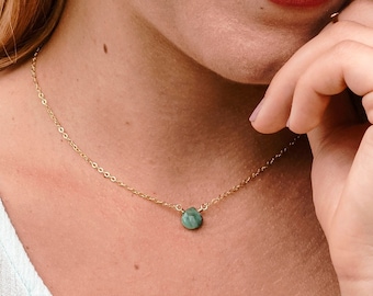 Pode incluir: Uma mulher vestindo uma blusa azul claro com um delicado colar de corrente dourada com um pequeno pingente redondo de pedra verde.