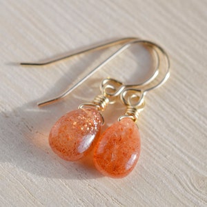May include: A pair of gold-colored drop earrings with teardrop-shaped, orange-colored sunstone beads. The beads have a sparkling, glittery appearance. The earrings are displayed on a light-colored wooden surface.