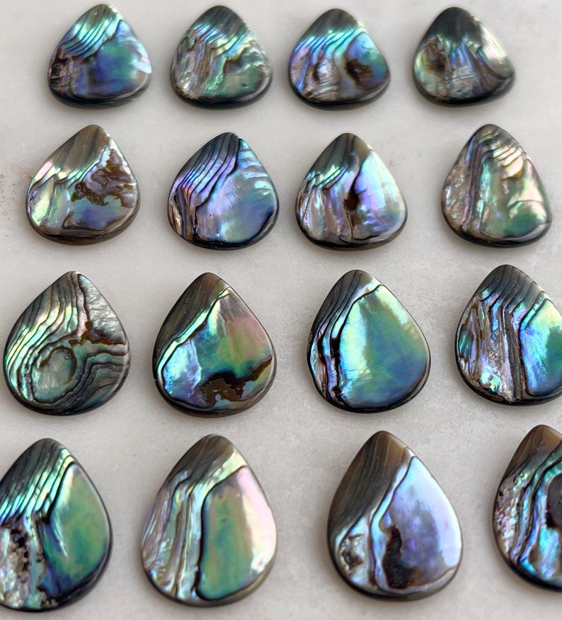 Puede incluir: Una colecci&oacute;n de 16 colgantes en forma de l&aacute;grima de concha de abul&oacute;n iridiscente. Las conchas son una mezcla de colores azules, verdes y morados con un acabado nacarado.