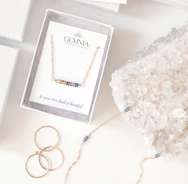 Puede incluir: Un collar de oro con una barra de piedras preciosas de colores y el texto "be your own kind of beautiful". El collar est&aacute; en una caja blanca con el texto "GEMNIA handcrafted jewelry".