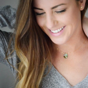 Puede incluir: Una mujer con el pelo largo y casta&ntilde;o claro lleva un su&eacute;ter gris y un collar de oro con un colgante de piedra verde e iridiscente. Est&aacute; sonriendo y mirando hacia abajo.