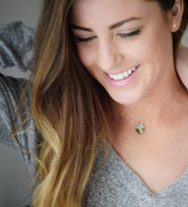Puede incluir: Una mujer con el pelo largo y casta&ntilde;o claro lleva un su&eacute;ter gris y un collar de oro con un colgante de piedra verde e iridiscente. Est&aacute; sonriendo y mirando hacia abajo.