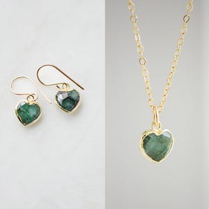 May include: Gold-toned heart-shaped earrings and a matching necklace. The earrings have a hook closure. The necklace features a delicate chain with a heart-shaped pendant. Both the earrings and pendant have a green gemstone center.