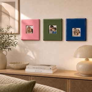 May include: Three square photo frames in glossy pink, olive green, and navy blue, each featuring a centred square aperture displaying a family photograph. The frames are mounted horizontally on a wall, showcasing a smooth, reflective finish.