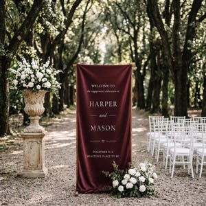 May include: A deep burgundy vertical fabric banner featuring white text that reads WELCOME TO the engagement celebration of HARPER and MASON TOGETHER IS A BEAUTIFUL PLACE TO BE, displayed on a wooden frame stand with white floral arrangements.