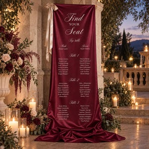 May include: A vertical burgundy satin fabric seating chart sign featuring the white text Find Your Seat, Top table, and lists of names organised by table numbers. The banner is adorned with a light beige ribbon bow at the top left corner.