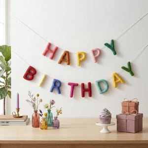 Può includere: Un colorato striscione in feltro con la scritta "HAPPY BIRTHDAY" è appeso a una parete bianca. Le lettere sono di vari colori, tra cui rosa, giallo e verde. Sotto, un tavolo mostra un cupcake, dei regali e piccoli vasi con fiori.