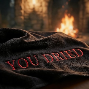 May include: A dark gray towel with the embroidered words "YOU DRIED" in red. The towel is folded and draped, with a blurred background featuring a fireplace. The towel appears soft and plush.