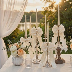 Puede incluir: Una colección de candelabros de plata y madera con velas cónicas blancas altas adornadas con lazos de cinta de satén color crema. Un pequeño jarrón de cerámica blanca lleno de flores blancas y color melocotón se sitúa junto a los candelabros.