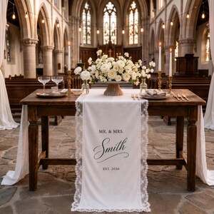 May include: A white fabric table runner with lace trim, printed with the text MR. & MRS. Smith EST. 2026. The runner is draped over a wooden table set with white floral arrangements, tall taper candles in gold holders, and glassware.