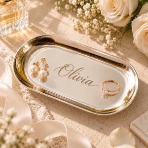May include: An oval silver-toned metal trinket tray featuring the name Olivia engraved in a cursive script. The tray holds a pair of gold-toned earrings with white pearls and a gold-toned ring with a clear stone setting.