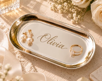 Könnte beinhalten: Eine ovale Schmuckschale aus silberfarbenem Metall mit dem eingravierten Namen Olivia in Schreibschrift. Die Schale enthält ein Paar goldfarbene Ohrringe mit weißen Perlen und einen goldfarbenen Ring mit einem klaren Stein.