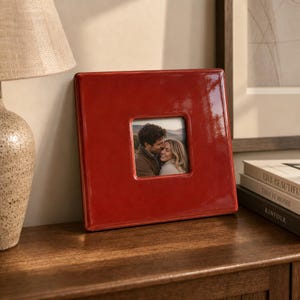 May include: A square, glossy red ceramic picture frame with a centered square opening. The frame holds a photograph of a person with dark hair embracing a person with blonde hair. The frame has a smooth, reflective surface and a thick border.