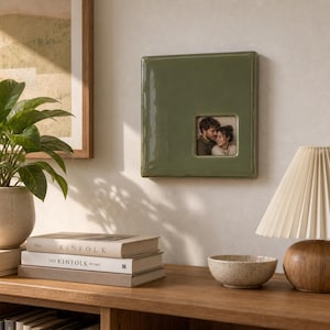 May include: A square, olive green ceramic wall-mounted photo frame featuring a glossy finish and a small, centered square cutout displaying a photograph of a couple embracing. The frame has a smooth, reflective surface and rounded edges.
