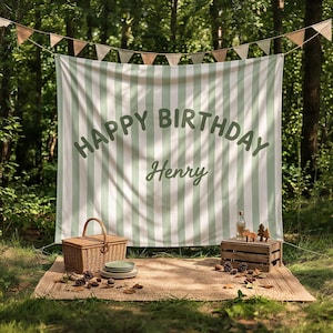 Op de afbeelding: Een grote rechthoekige stoffen achtergrond met verticale saliegroene en witte strepen met de tekst HAPPY BIRTHDAY Henry in donkergroene letters, opgehangen aan een slinger van driehoekige vlaggetjes boven een gevlochten picknickmand en krat.