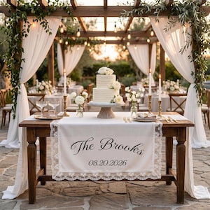 May include: A two-tier white wedding cake with horizontal ridges and white floral accents sits on a wooden table. A white runner with lace trim features the text The Brooks and the date 08.20.2026. The table includes gold candlesticks and glassware.