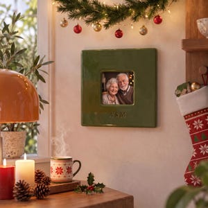 May include: A square, olive green ceramic picture frame featuring a glossy finish and the engraved letters A & M at the bottom. The frame holds a photograph of an older couple and is mounted on a wall decorated with holiday lights.