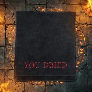 Könnte beinhalten: Ein schwarzes Badetuch mit dem gestickten Text "YOU DRIED" in Rot. Das Handtuch ist rechteckig und scheint aus einem weichen Material zu sein. Der Hintergrund zeigt eine steinartige Oberfläche mit leuchtenden Akzenten.