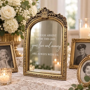 May include: An arched, ornate gold-toned metal frame mirror featuring the white printed text: THOUGH ABSENT FROM THIS DAY, your love and memory ARE ALWAYS WITH US. The frame has intricate floral and scrollwork detailing along the top edge.