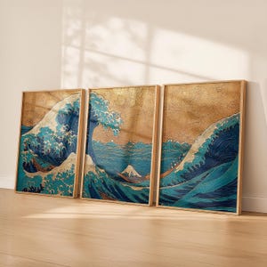 Puede incluir: Tres impresiones artísticas enmarcadas que representan una ola oceánica estilizada en tonos azules y blancos, con un fondo dorado. La obra de arte recuerda a las estampas japonesas, con una montaña visible en la distancia. Los marcos son de color madera clara.