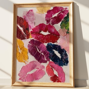 May include: A framed artwork featuring a collection of colorful lipstick kiss marks. The kisses are painted in various shades of red, pink, purple, yellow, and blue, creating a vibrant and artistic display. The artwork is framed in a light-colored wooden frame.
