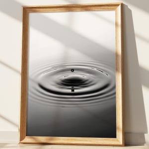 May include: A framed black and white photograph of a water droplet creating ripples. The image is in a wooden frame, and the water's surface is dark with concentric circles emanating from the point of impact. The photograph captures a moment of stillness and movement.