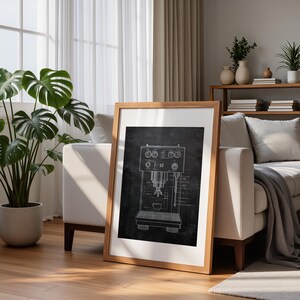 May include: A framed print featuring a detailed blueprint of an espresso machine. The artwork is rendered in white lines against a dark background, encased in a light brown wooden frame. The print is leaning against a white sofa in a well-lit room.