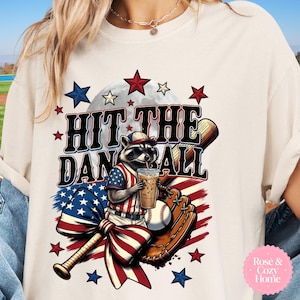 Puede incluir: Camiseta color crema con un mapache con gafas de sol y uniforme de béisbol, sosteniendo una bebida. La camiseta tiene el texto "HIT THE DAN BALL" con estrellas y un diseño de bate, guante y pelota de béisbol. También hay un lazo con la bandera estadounidense.
