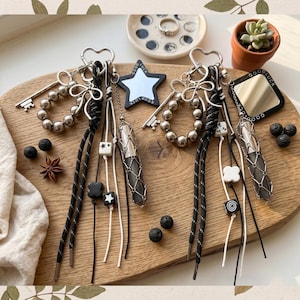 Peut inclure: Deux porte-clés avec des accessoires argentés, des perles et des breloques décoratives. Chaque porte-clés comprend un pendentif en cristal, des cordons noirs et blancs et un charme en forme d'étoile ou de miroir avec l'inscription "Good Luck".