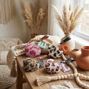 May include: A selection of fluffy heart-shaped keyrings in shades of pink, leopard print, and brown. Each keyring has a leather strap and a letter charm. The keyrings are arranged on a wooden table, alongside decorative items such as vases and beads.